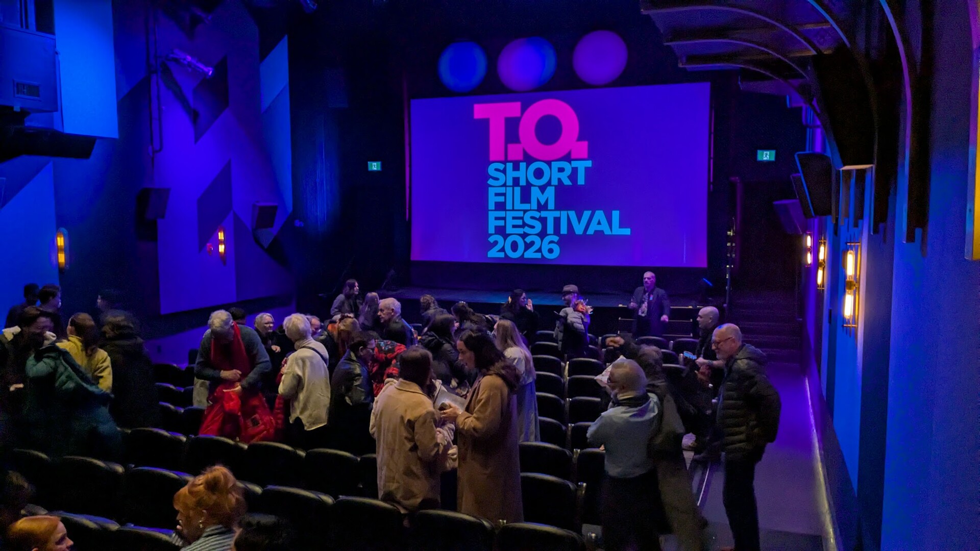 Audience members stand in the aisles of a cinema auditorium while the T.O Short Film Festival 2026 title card glows on the screen.
